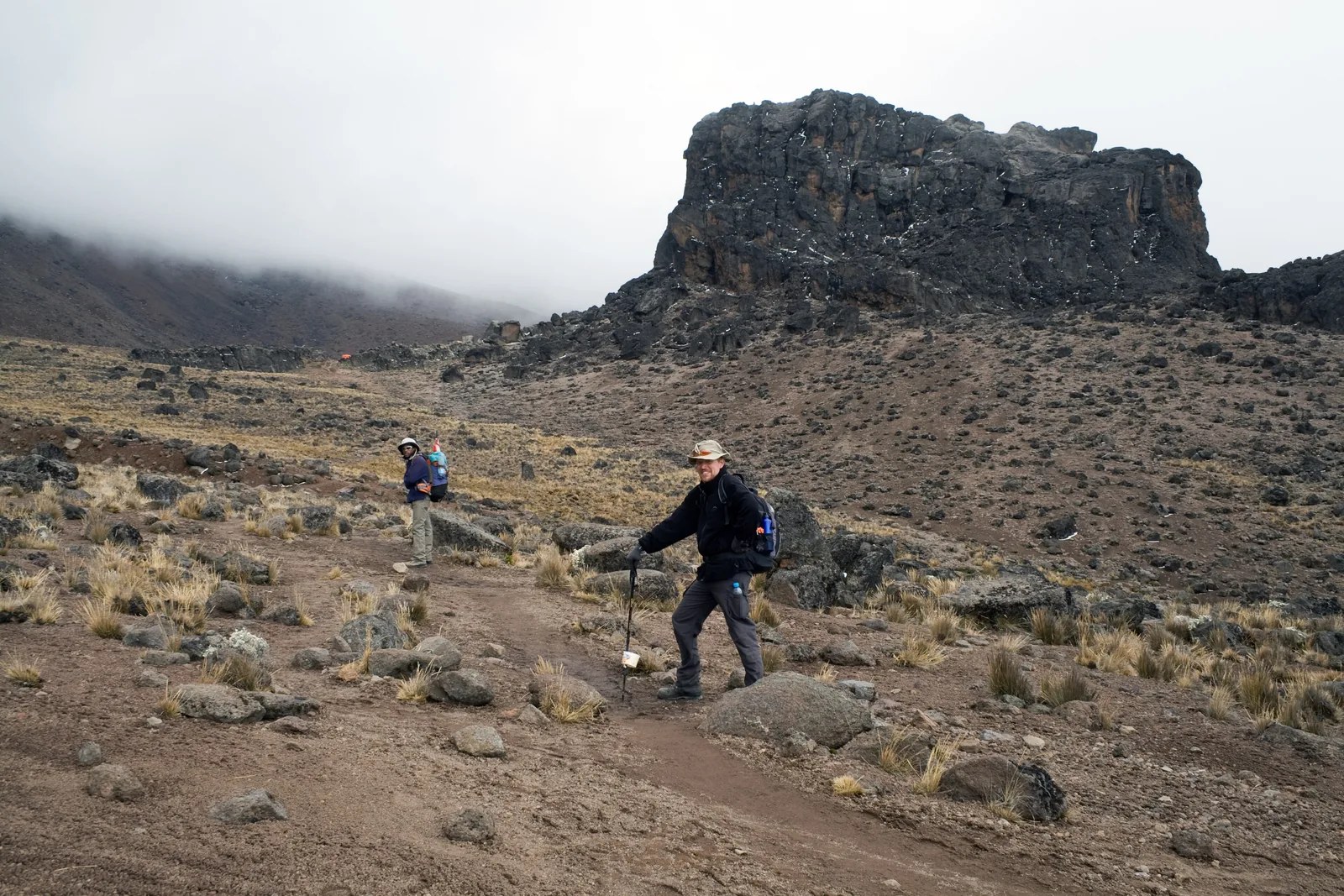 Hikers with poles in front of the Lava Tower in Kilimanjaro National Park, a dark rock tower above scree and cloud