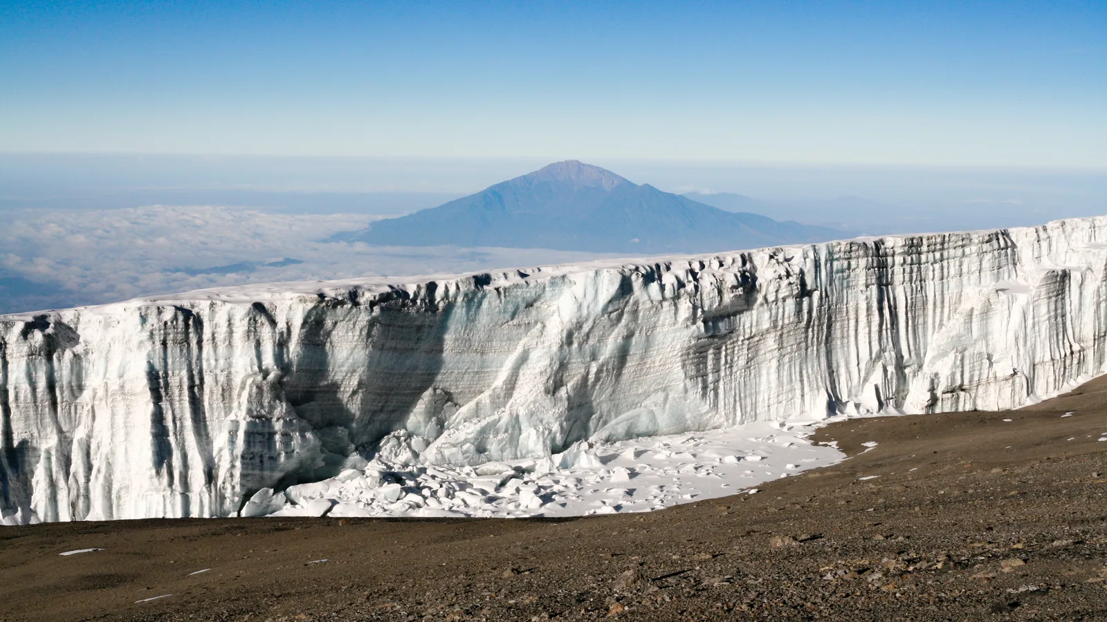 A vertical glacier wall near Uhuru Peak in Kilimanjaro National Park, with Mount Meru above the clouds in the distance
