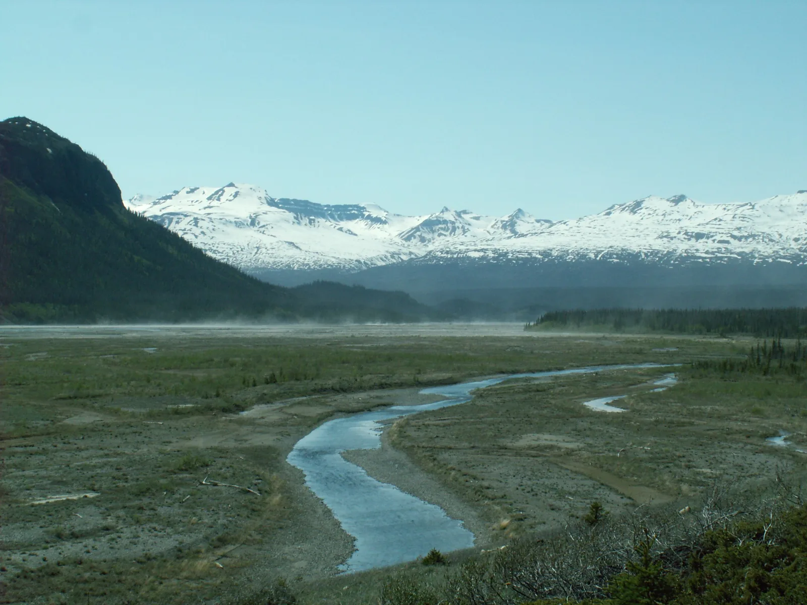 The braided Alsek River in Kluane National Park across a wide gravel plain, behind it a snow-covered mountain range in haze