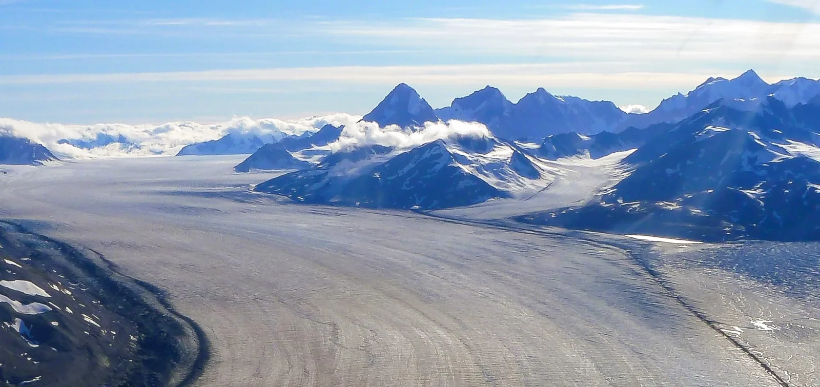The Lowell Glacier in Kluane National Park runs as a broad ice stream through the picture, behind it Ulu Mountain and other peaks above a layer of cloud