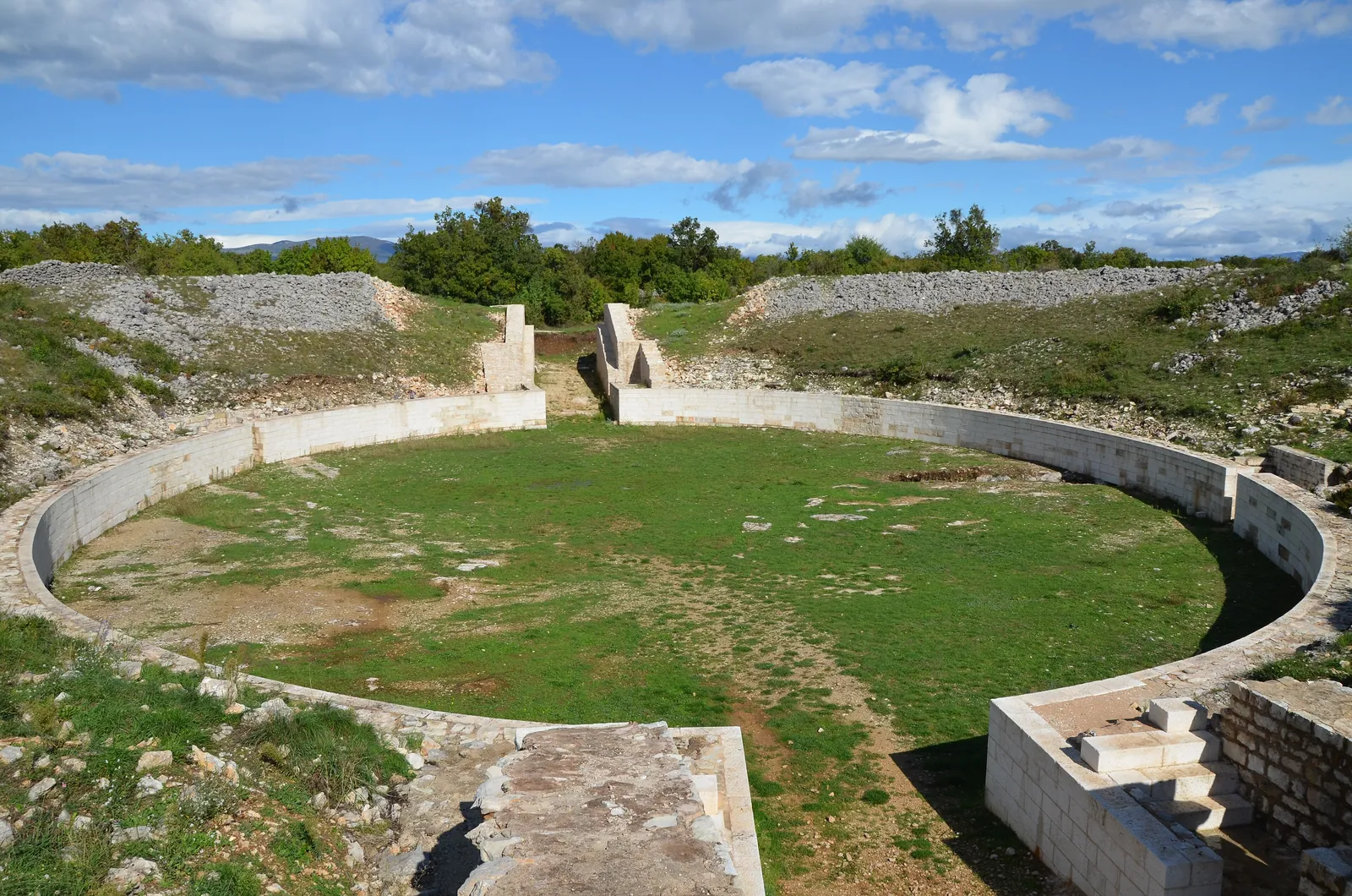 The oval arena of the Roman amphitheatre at Burnum in Krka National Park with pale stone walls, a grass floor and two entrances under a blue sky with clouds