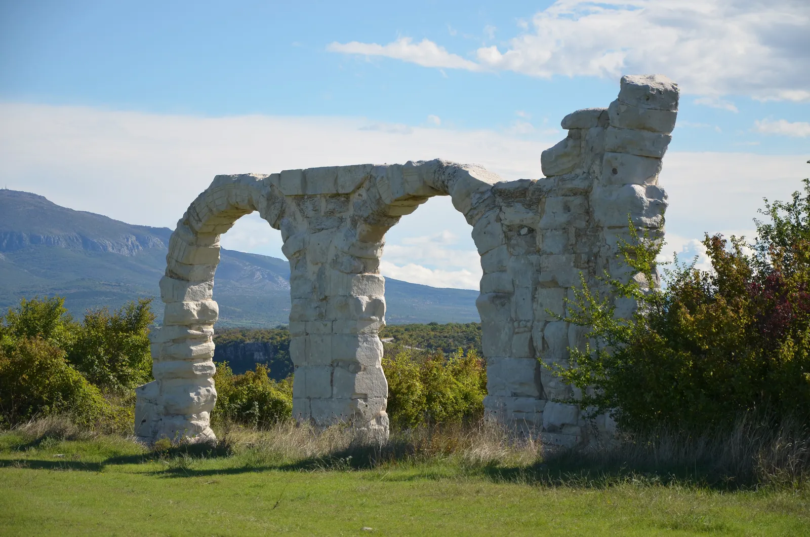 Two tall Roman arches of pale limestone stand in Krka National Park in a meadow, with scrub and a wooded ridge under a blue sky behind