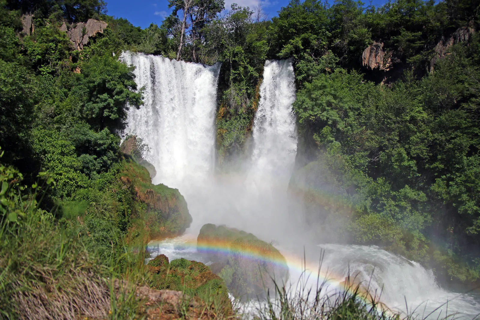 Two mighty white waterfalls plunge in Krka National Park down between green scrub, with a rainbow in the spray above mossy rocks below