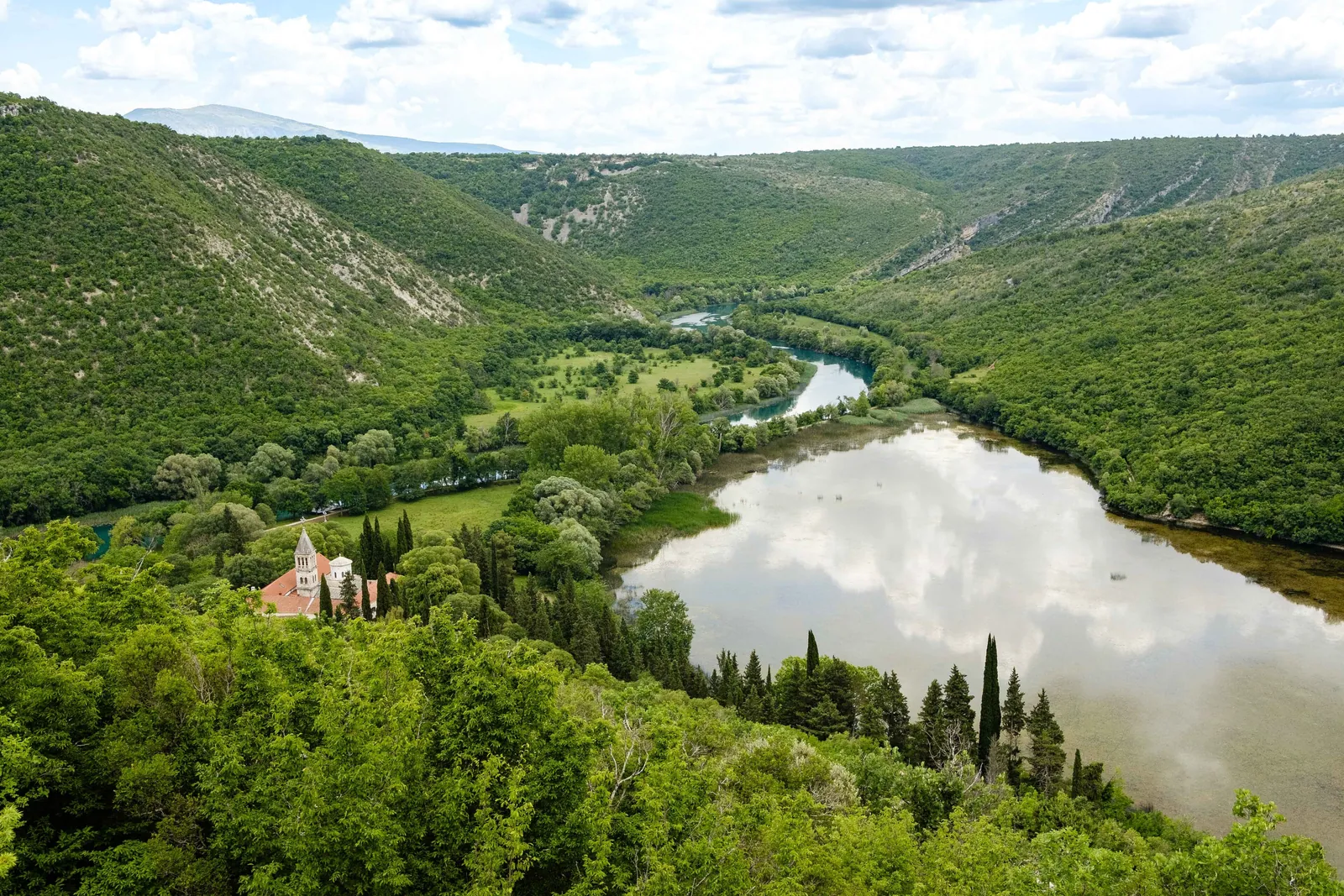 View from above into the green Krka valley in Krka National Park: the monastery with its bell tower between cypresses on the left, the broad, mirror-like river on the right