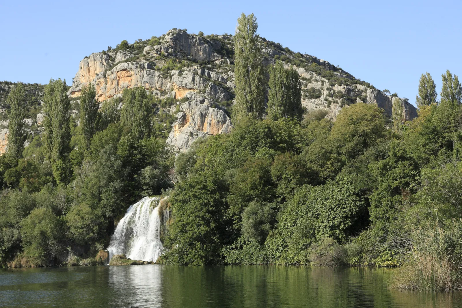 A white waterfall plunges in Krka National Park between dense greenery into a calm lake, with tall poplars and pale limestone rocks under a blue sky behind
