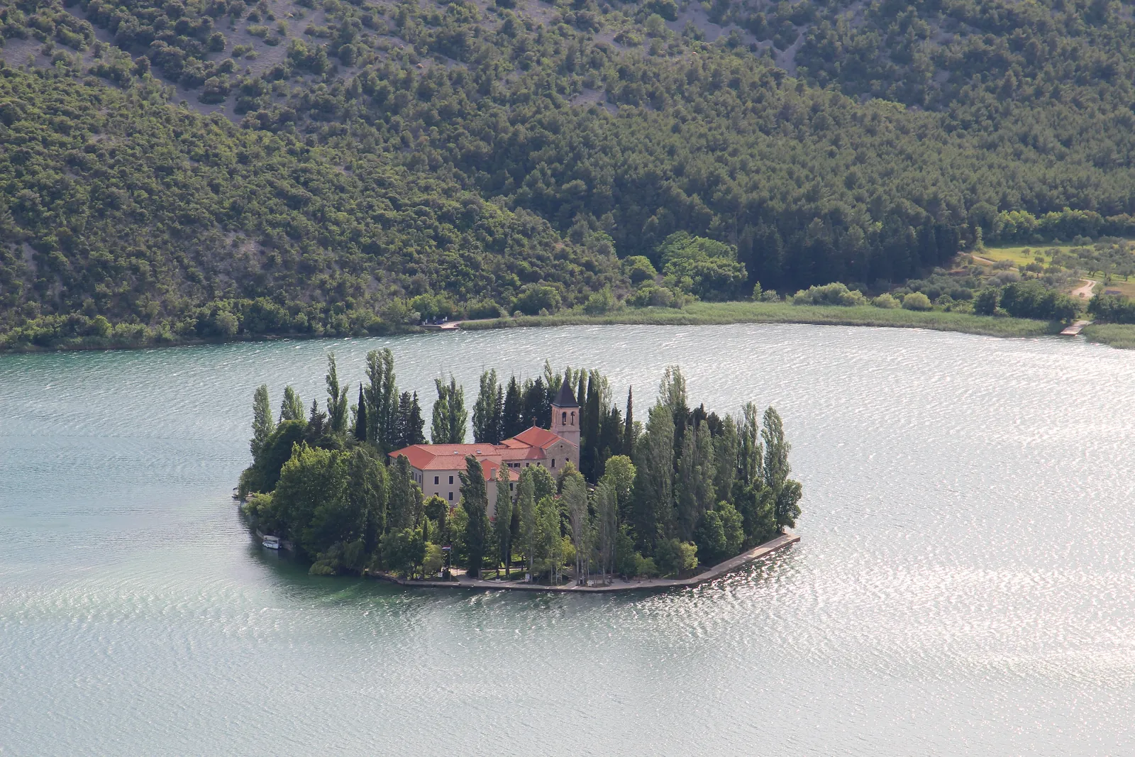 The small island of Visovac in Krka National Park with its monastery building, red tiled roof and bell tower between tall poplars and cypresses, surrounded by silvery water