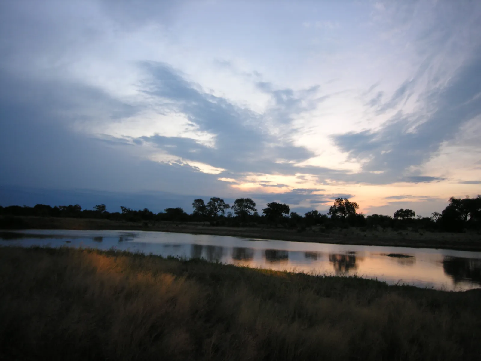 Dusk over a still reservoir, a dark line of trees on the shore, veils of cloud above in the last light