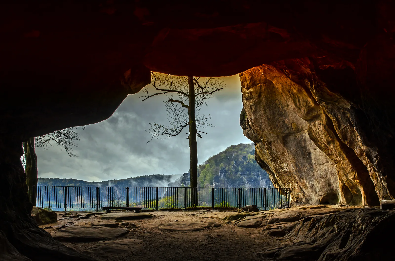 View through the Kuhstall rock arch in Saxon Switzerland National Park to the wooded Affensteine, with the viewpoint railing in the foreground