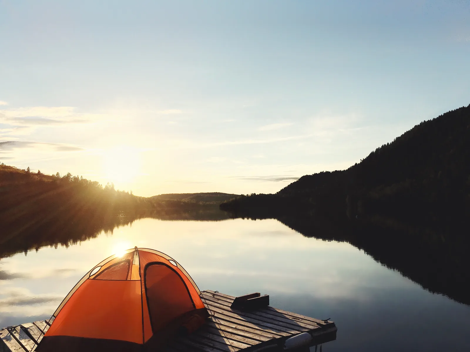 An orange tent stands on a wooden platform on the lakeshore in La Mauricie National Park, the sun rising behind the forested hills and reflected in the still water