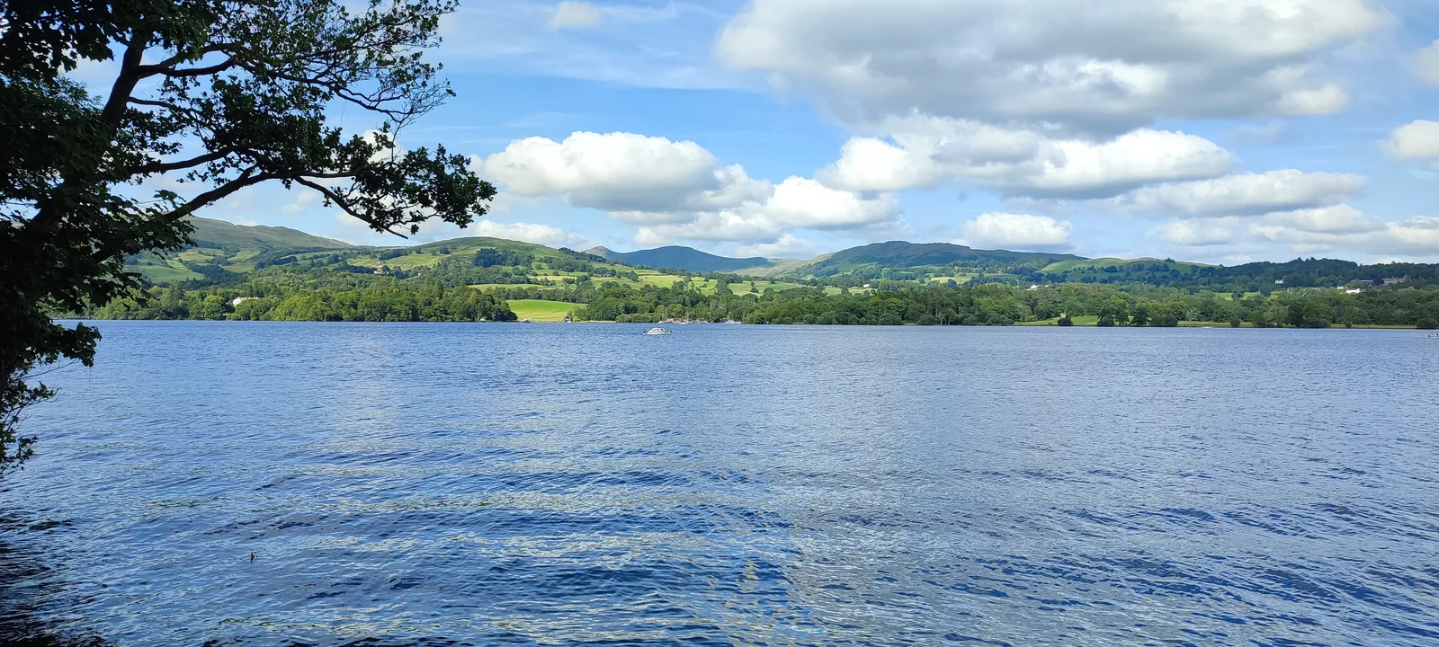Windermere in the Lake District National Park under a bright cloudy sky, trees on the shore and low hills behind