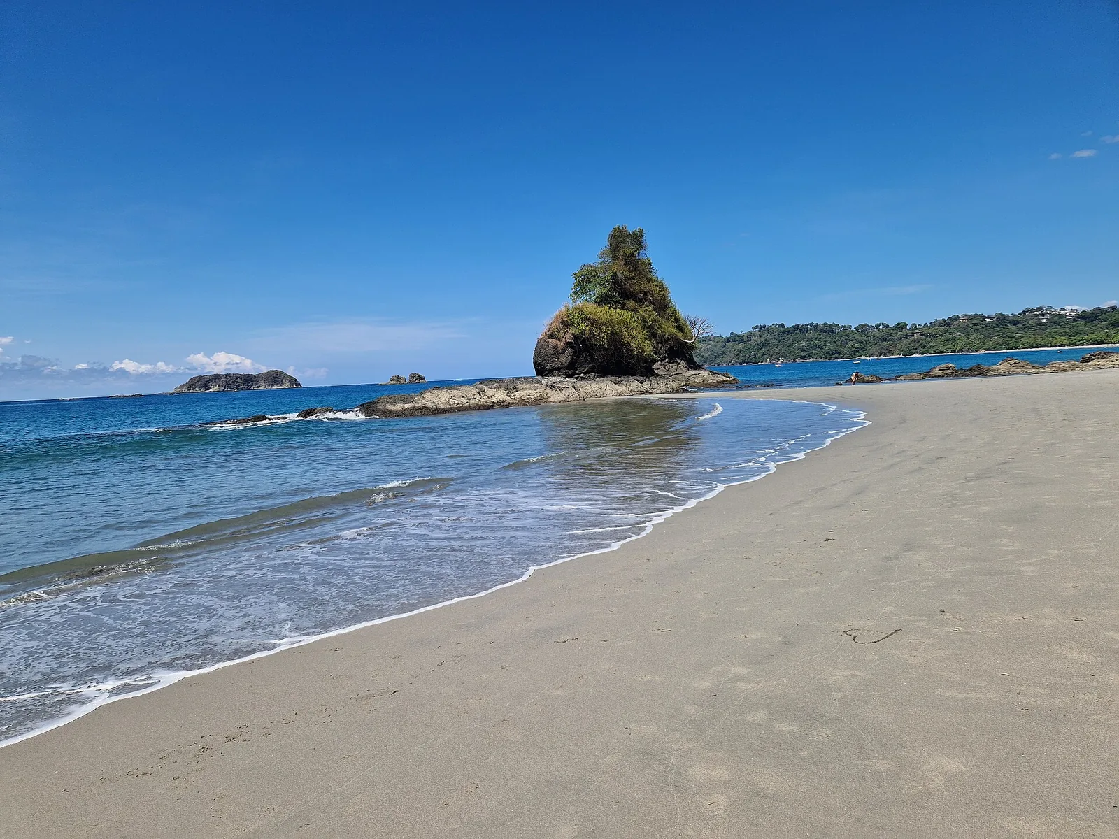 White sand beach in Manuel Antonio National Park with a forested rock island in the turquoise Pacific under a blue sky