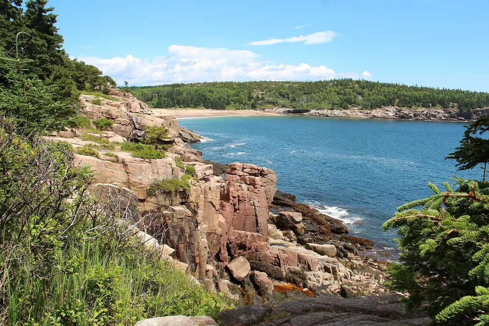 Pink granite cliffs at Otter Cliff in Acadia National Park, with a bay, a sandy beach and a wooded shore behind