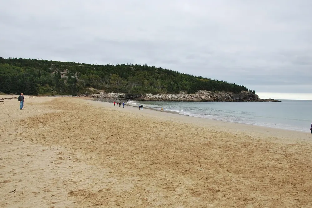 Sand Beach near the fee station in Acadia National Park: a wide sandy beach between a wooded headland and the sea, a few people walking