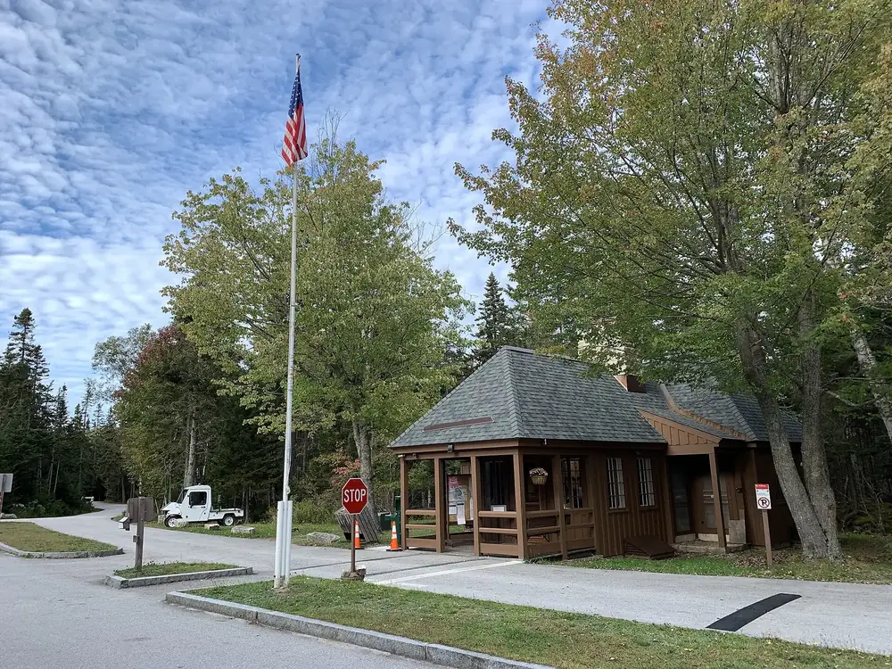 Entrance to Seawall Campground in Acadia National Park, with a wooden fee booth, a flagpole and tall deciduous and conifer trees