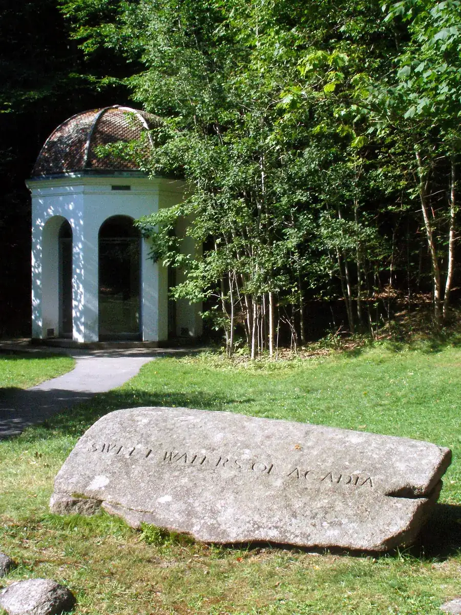 Small white springhouse in Acadia National Park over the Sieur de Monts spring, with a granite block with a carved inscription in front