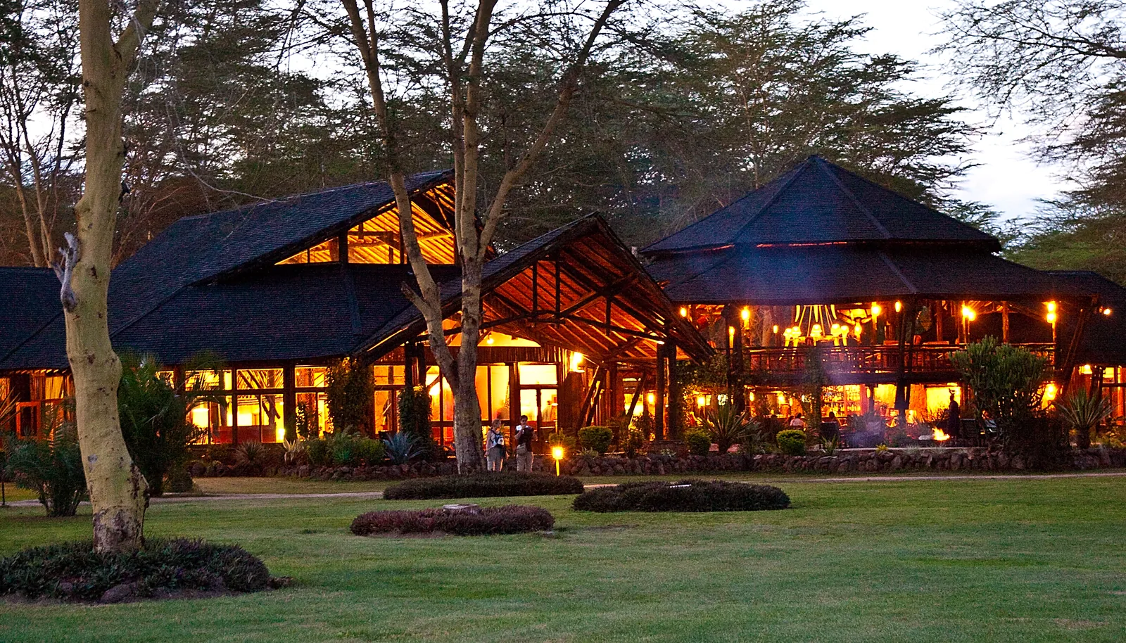 The lit main building of Ol Tukai Lodge in Amboseli National Park in the evening, with tall pointed roofs between acacias