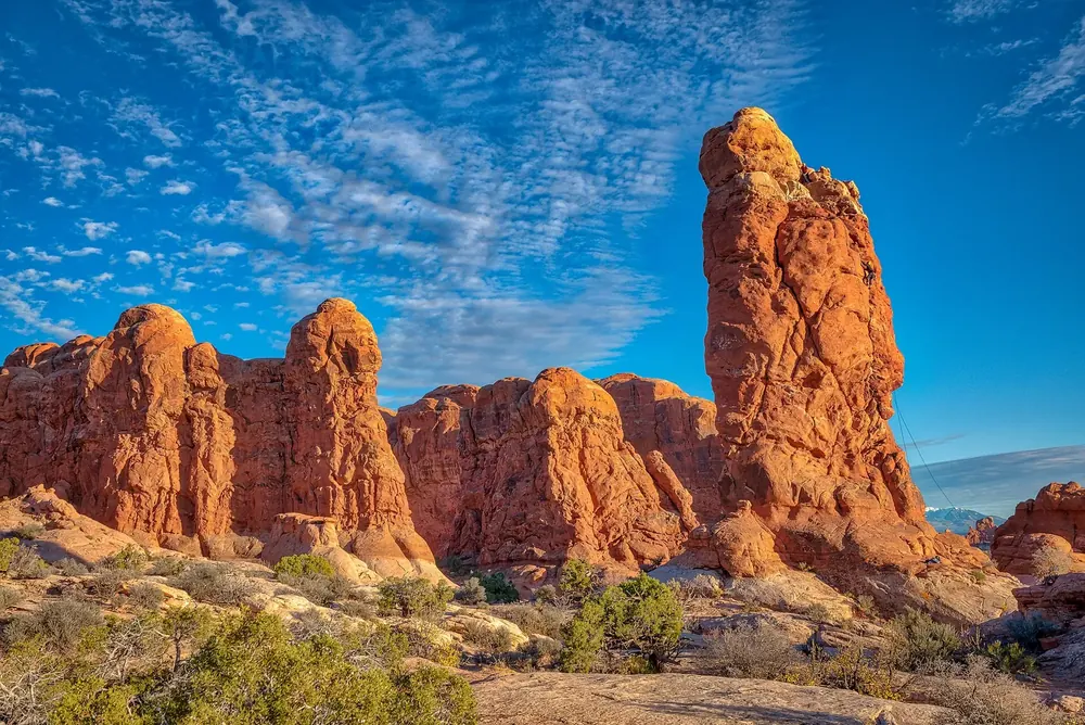 Garden of Eden in Arches National Park: red sandstone columns and rock towers under a blue sky with wispy clouds, low desert vegetation in front