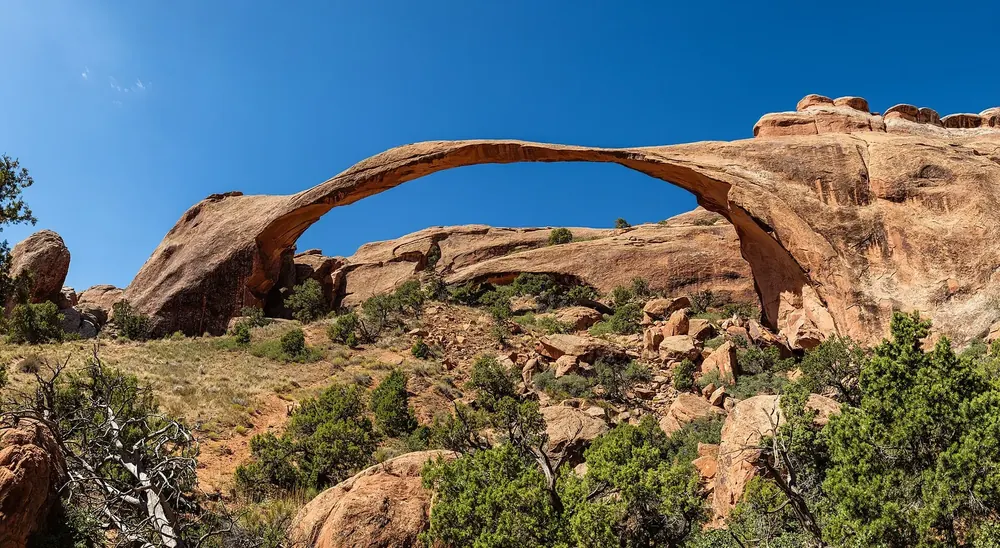 Landscape Arch in Arches National Park: an extremely long, thin sandstone arch above boulders and green juniper under a deep blue sky