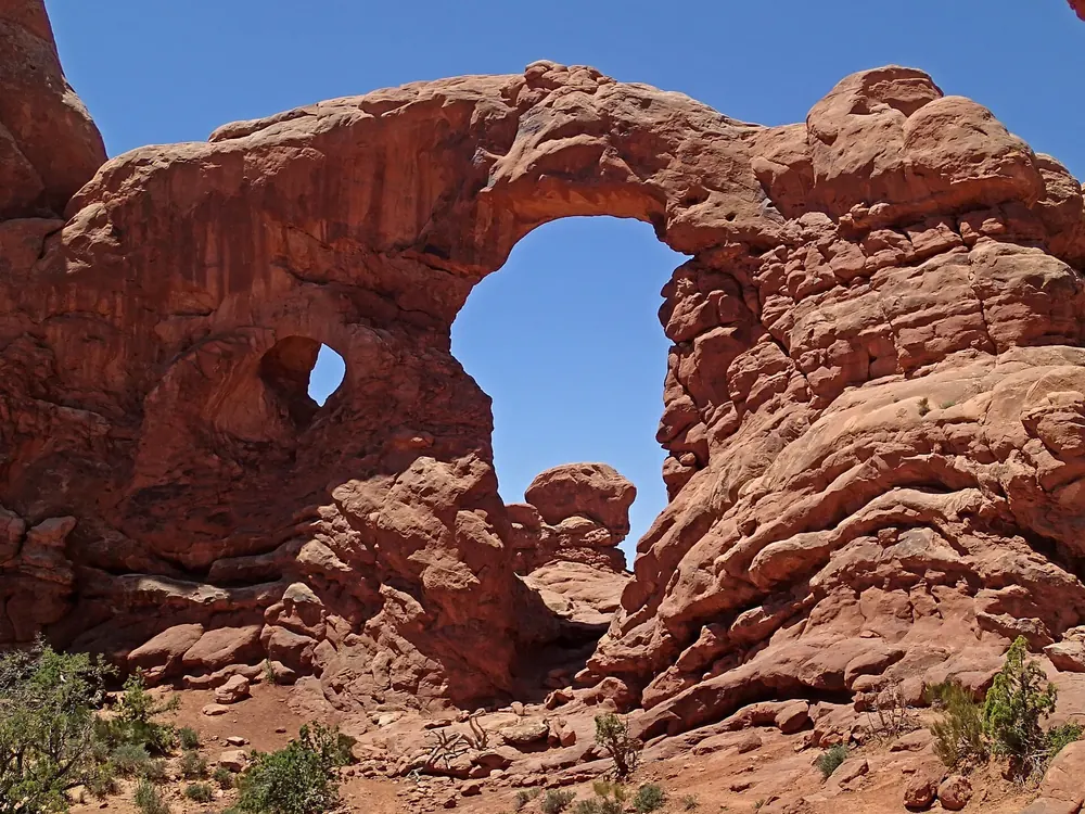 Turret Arch in Arches National Park: a sandstone arch with a large and a small window, beside a tower-like rock pillar under a blue sky