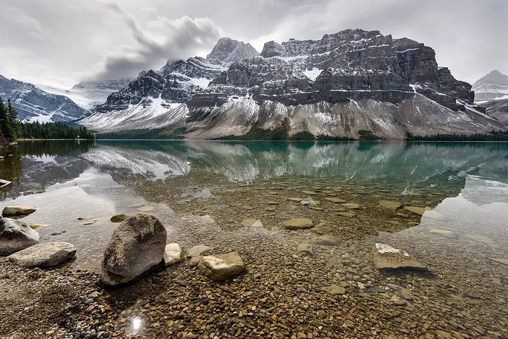 Bow Lake in Banff National Park reflecting snow-covered rock peaks, stones on the shore in the clear water under clouds