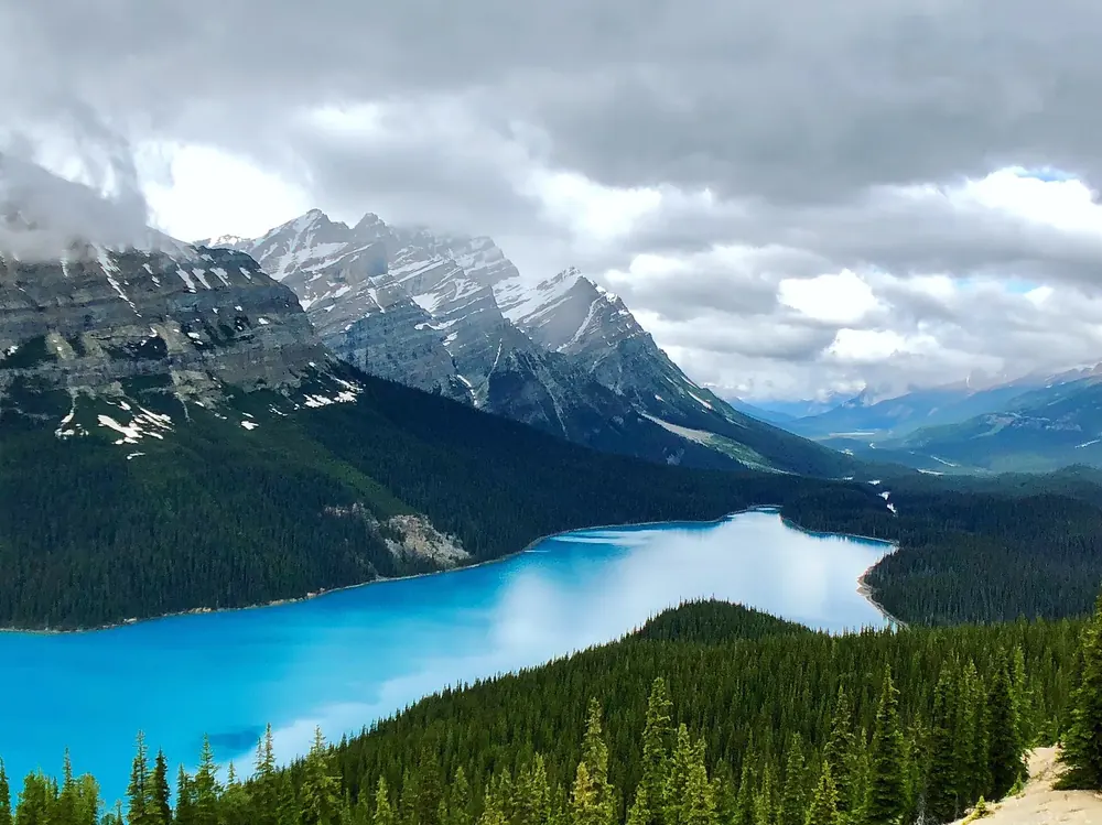 View from Bow Summit in Banff National Park of bright turquoise Peyto Lake, a forested valley and cloud-covered peaks
