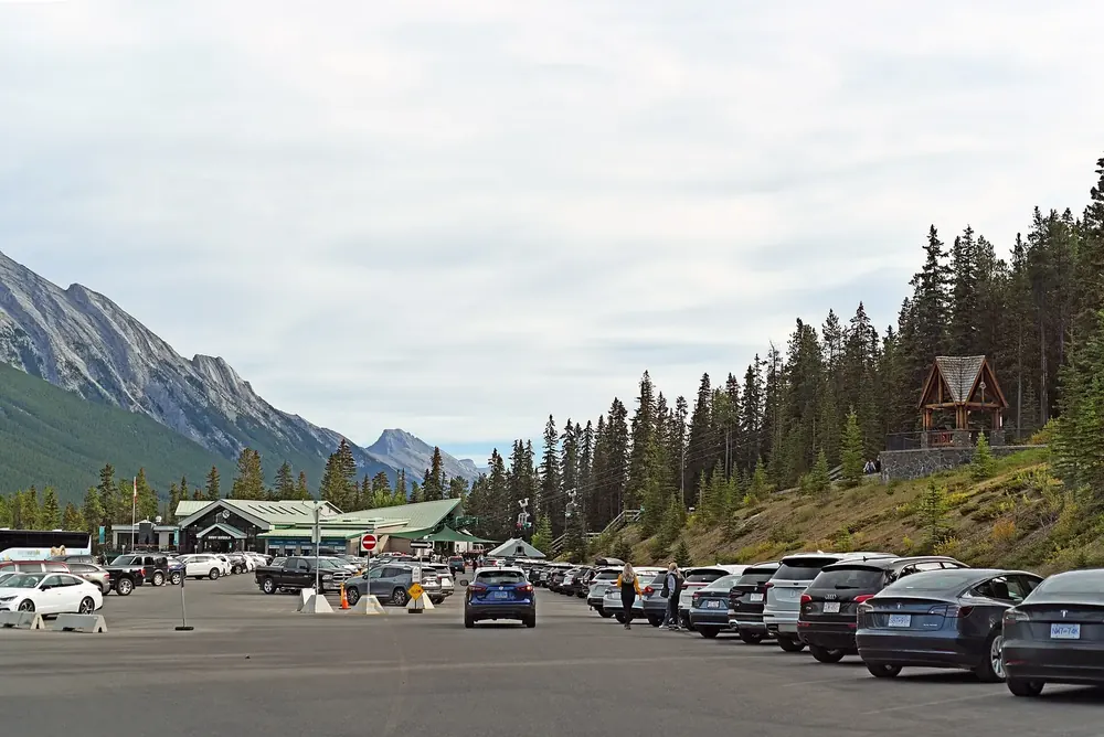 Parking lot at the Banff Gondola base station in Banff National Park, lined with conifer forest, steep grey mountainsides behind.