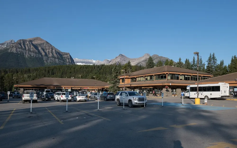 Samson Mall in the village of Lake Louise in Banff National Park next to the visitor centre: wooden buildings, parking lot and shuttle bus below mountains.
