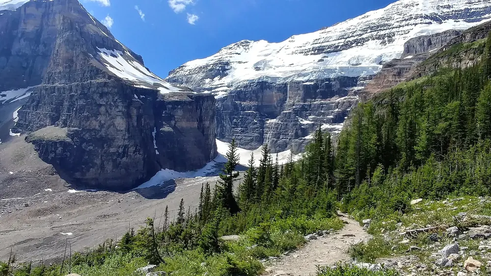 Trail through the Plain of Six Glaciers in Banff National Park: scree slopes, conifer forest and glaciated rock faces under a blue sky.