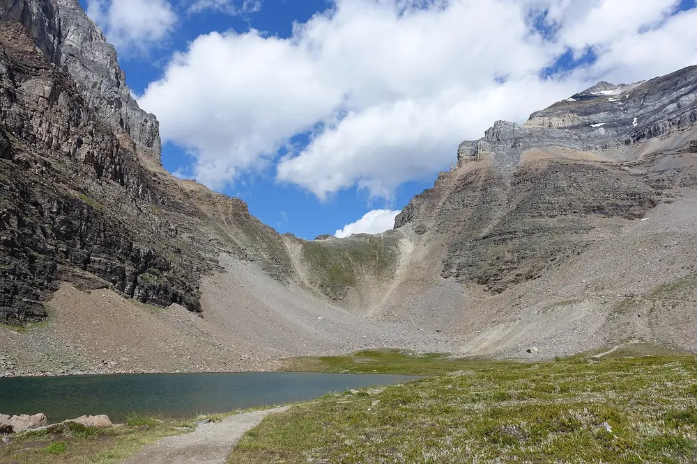 Sentinel Pass in Banff National Park: scree saddle between steep rock walls, a small mountain lake and a trail over alpine meadows in front.
