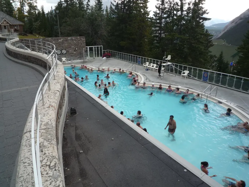 Outdoor pool of the Banff Upper Hot Springs in Banff National Park with bathers in the turquoise thermal water, conifer forest on the slope behind.