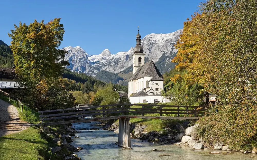 Parish church of St. Sebastian in Ramsau with a wooden bridge over the stream, autumn leaves and snow-covered peaks behind