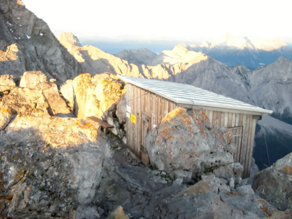Small wooden shelter hut on the rocky Hocheck summit of the Watzmann in Berchtesgaden National Park, mountain ranges behind in evening light