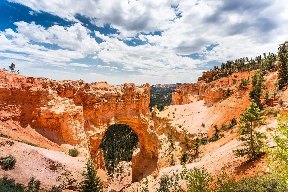 Natural Bridge in Bryce Canyon National Park: a red-orange rock arch over wooded ground, surrounded by eroded slopes under cumulus clouds