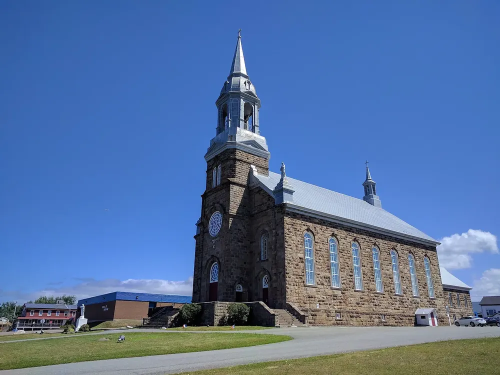 Large stone church in Chéticamp with a tall, pointed spire under a deep blue sky
