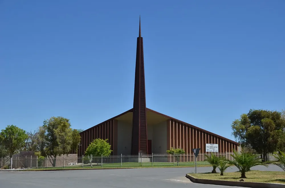 Church in Outjo with a tall, pointed tower and steep roof, trees and palms in front under a blue sky