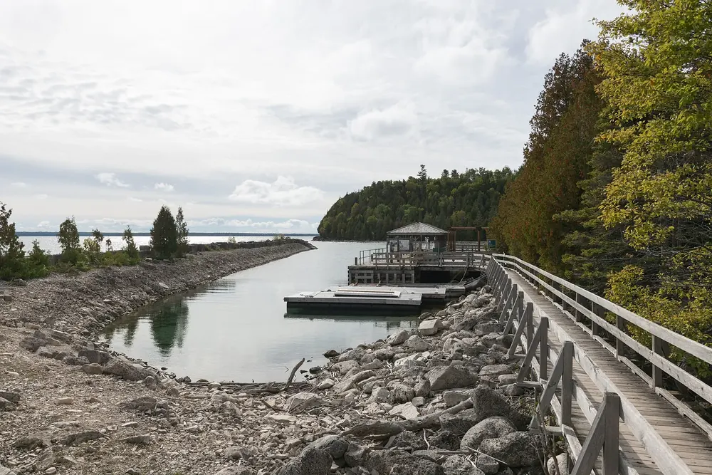 Boat dock on Flowerpot Island: wooden pier and gravel shore on a calm bay, wooded rocky coast behind