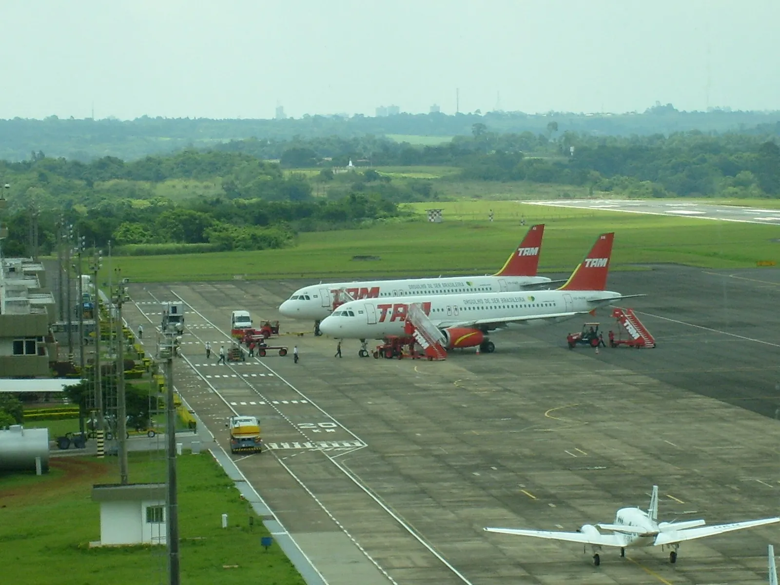 Apron of Foz do Iguaçu airport from above with two TAM airliners and a light aircraft, green hills beyond