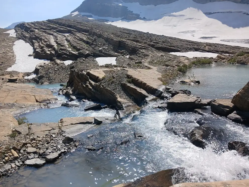 Meltwater from Grinnell Glacier in Glacier National Park, Montana, pours over layered limestone, with snowfields and ice behind
