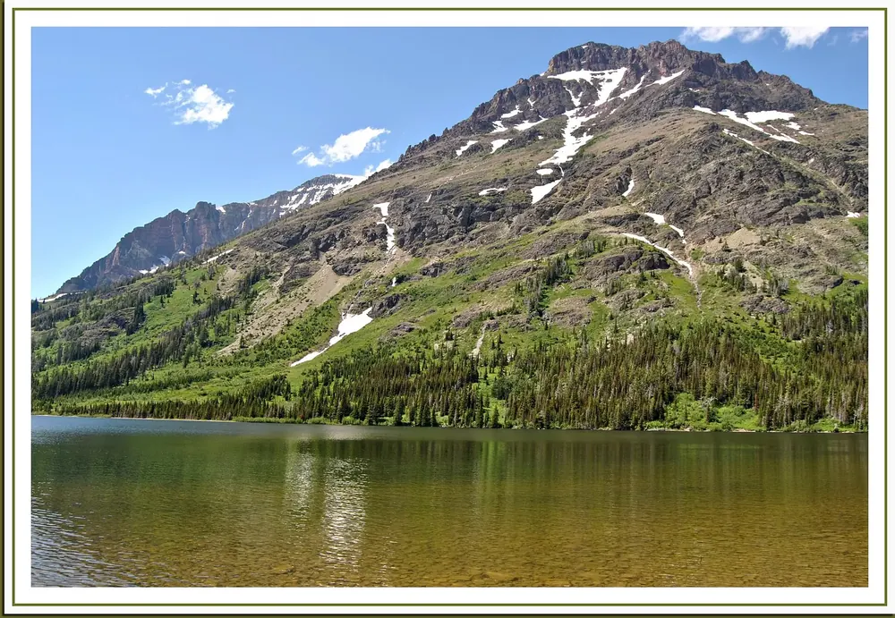 Two Medicine Lake in Glacier National Park, Montana: a clear mountain lake in front of a green mountainside with patches of snow under a blue sky