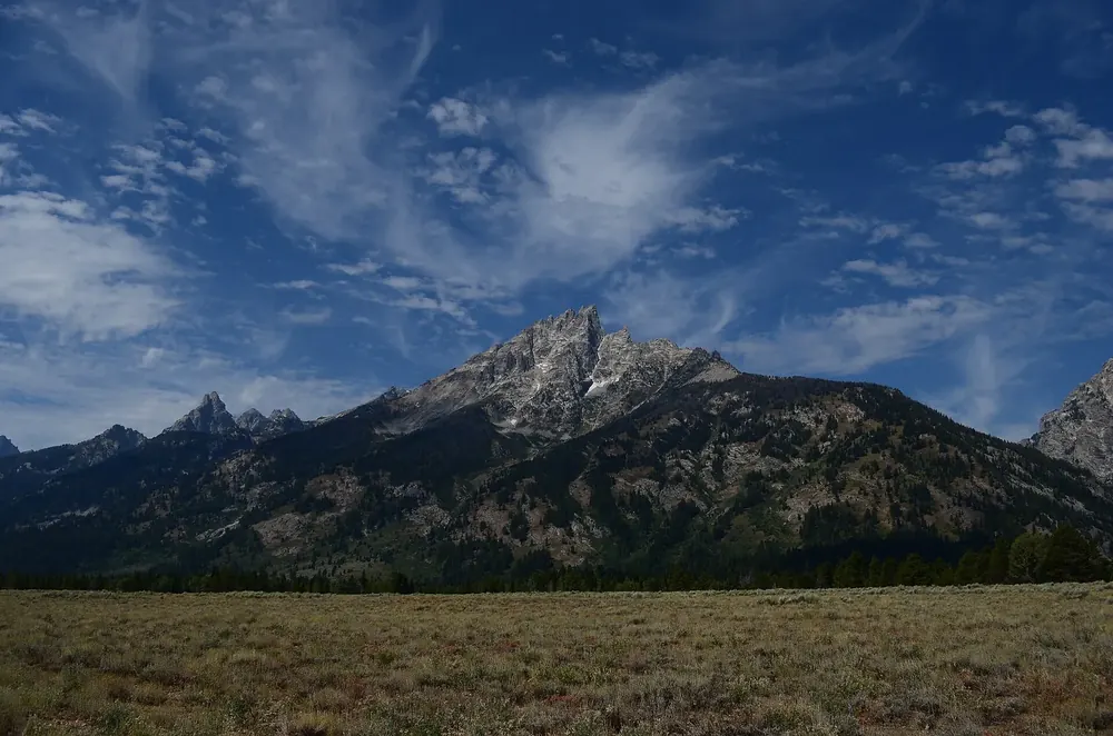 Grand Teton, the highest peak in Grand Teton National Park, rising above dark forested slopes and a sagebrush plain, with wispy clouds in the sky