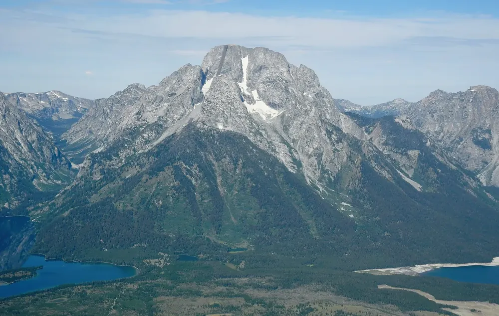 Aerial view of Mount Moran in Grand Teton National Park from the east: a massive granite peak with snowfields above forested slopes and a lake