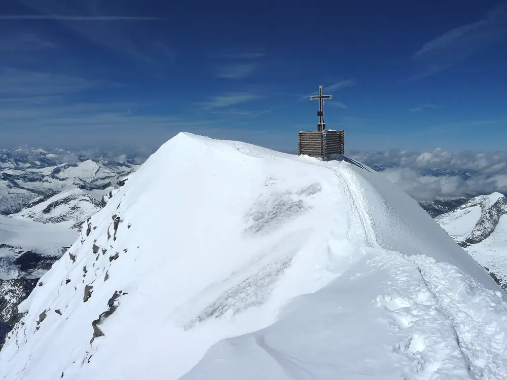 Summit of the Grossvenediger in Hohe Tauern National Park: snowy firn ridge with a summit cross above a sea of glaciated mountains