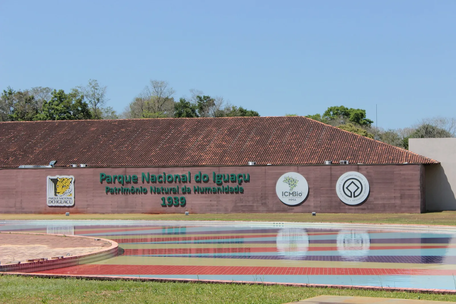 Entrance building of Iguaçu National Park with a tiled roof and the lettering “Parque Nacional do Iguaçu – Patrimônio Natural da Humanidade 1939”, a colourful pool in front