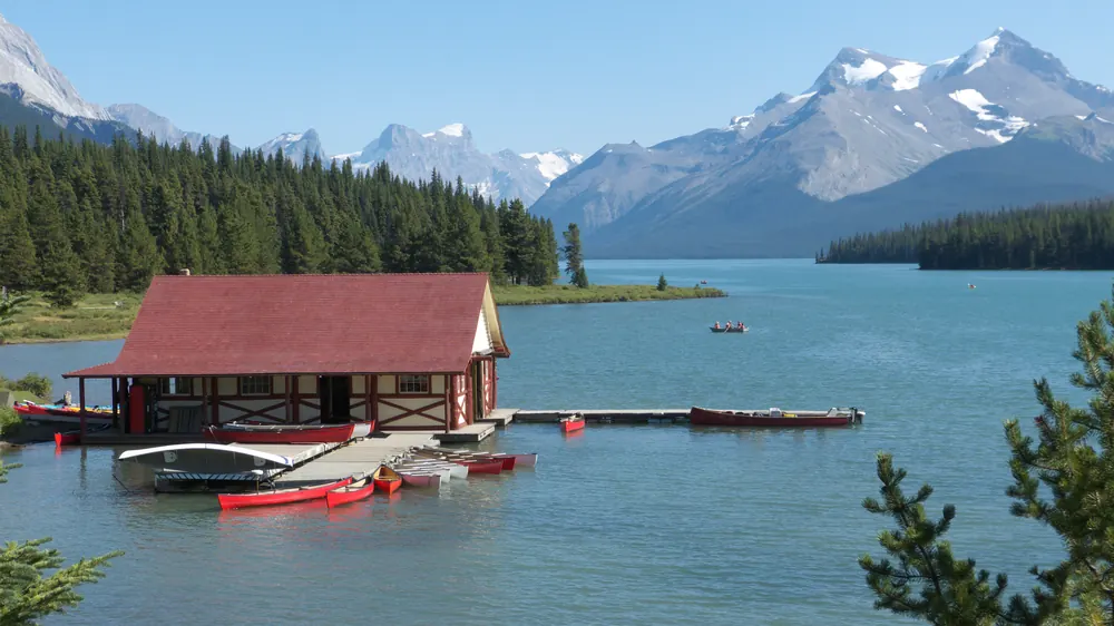 Red boathouse on Maligne Lake in Jasper National Park with a dock and red canoes, conifer forest and snow-capped Rocky Mountain peaks behind
