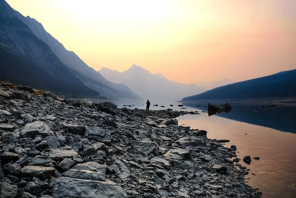 Medicine Lake in Jasper National Park in morning light: rocky shore, still lake and hazy mountain ranges in the smoke of nearby wildfires