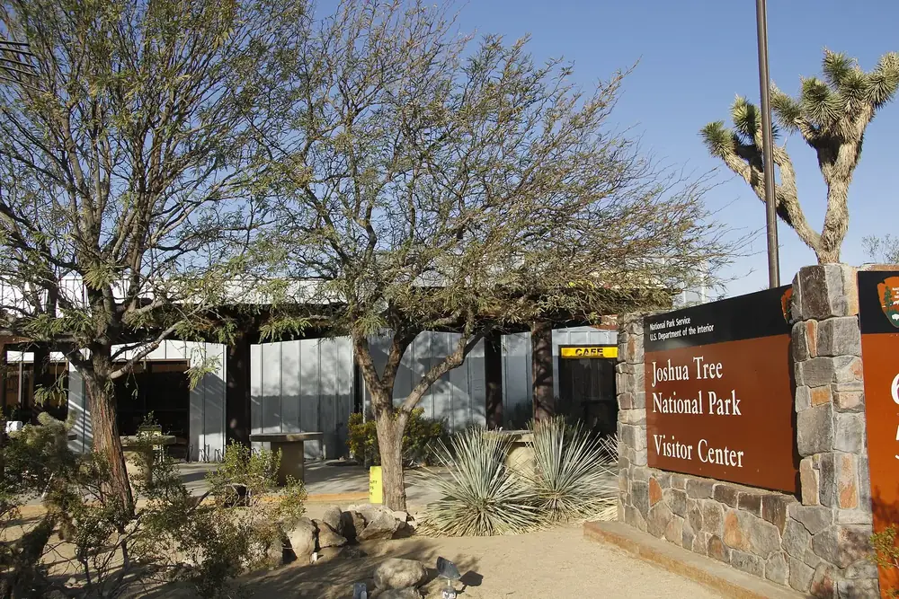 Joshua Tree Visitor Center in the town of Joshua Tree: entrance with a stone wall and park sign, desert plants and a Joshua tree in front