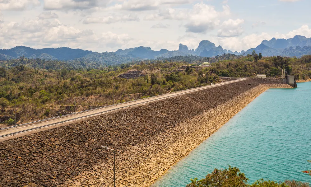 Rockfill wall of the Ratchaprapha Dam with a road, turquoise Cheow Lan Lake on the right, wooded karst mountains behind