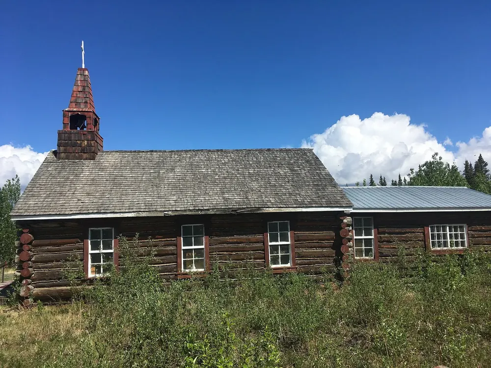 Old log church in Burwash Landing with a small wooden bell tower on a meadow under a blue sky