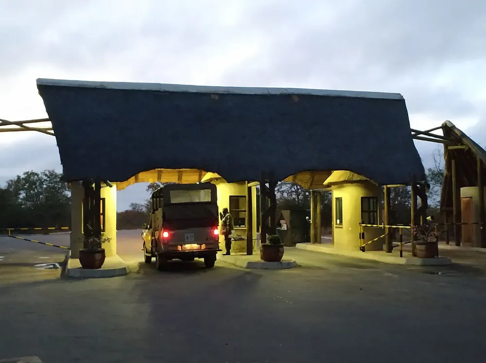 Phalaborwa Gate at sunrise: lit gate building with a large thatched roof over the access road, a safari vehicle below