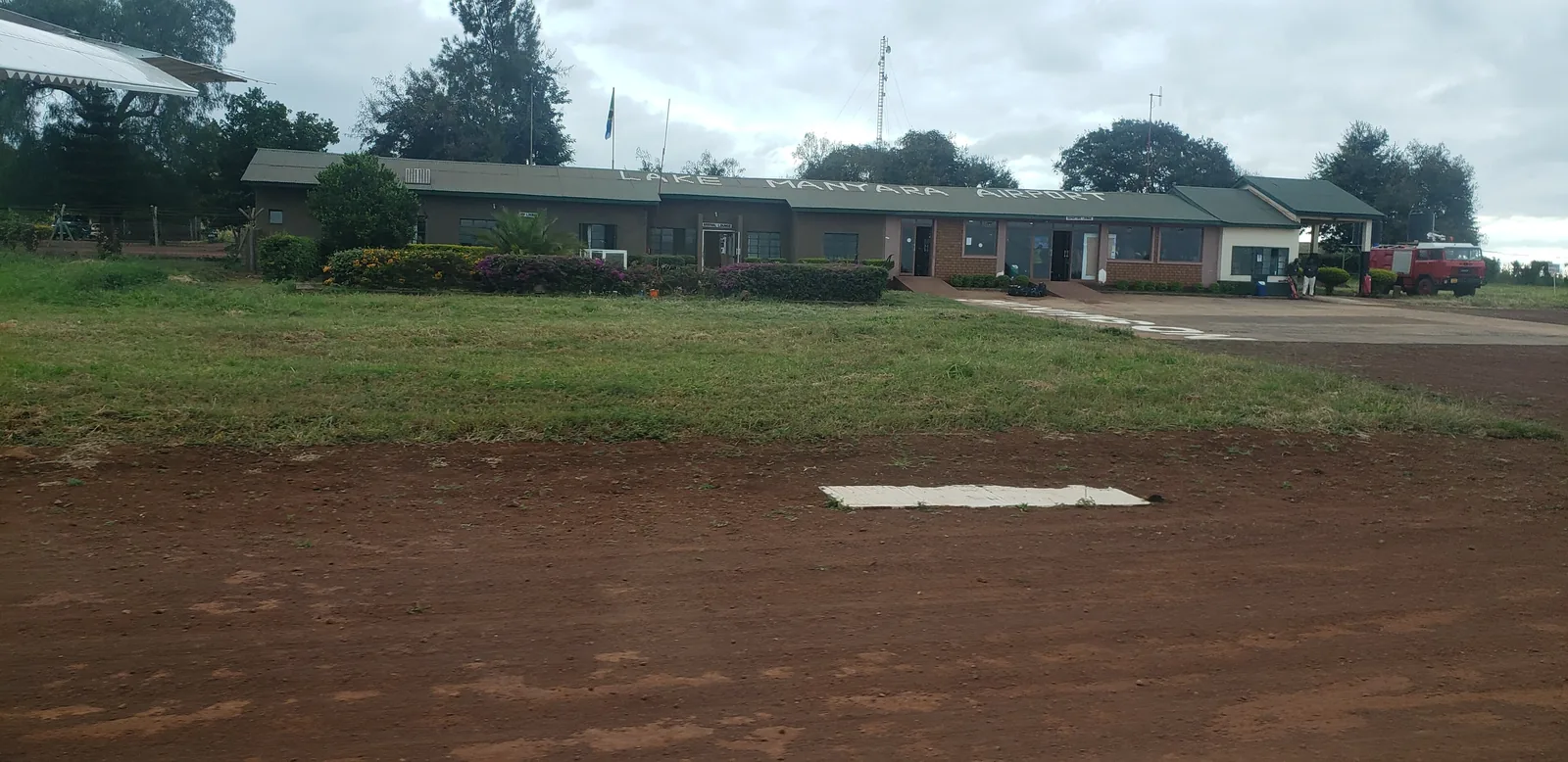 Low terminal building of Lake Manyara Airport behind a lawn and the red apron, outside the national park
